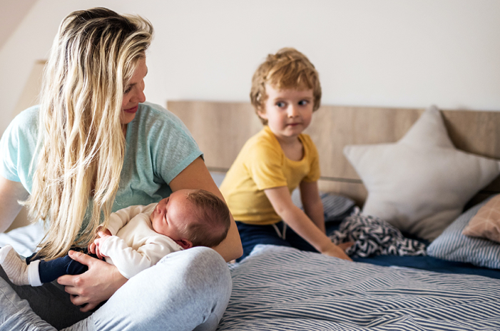 Woman sitting on bed holding infant while another young child looks on in background