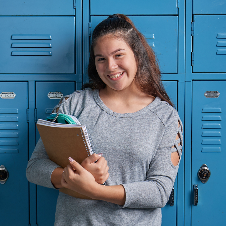 Adolescent girl holding notebook and smiling in front of lockers