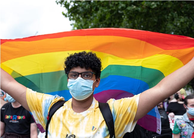 Youth Wearing Glasses and a Mask Holding Pride Flag Over Their Head